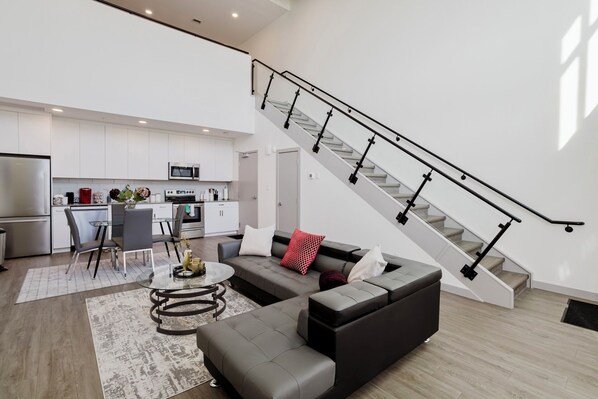 A modern apartment with a kitchen, dining area, and living room. It features a staircase with a black railing and a large window providing natural light. - Luxe Loft, King bed, 10 mins from Dt (Edmonton)