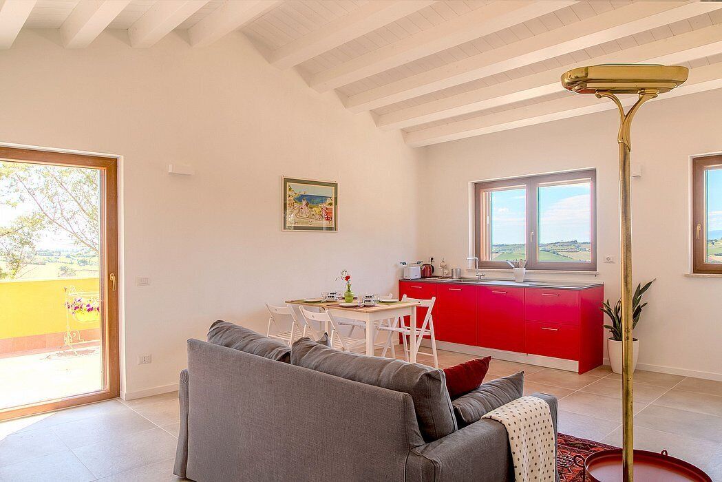 A modern living room with a grey sofa, a dining table, and a red kitchen area with a window.