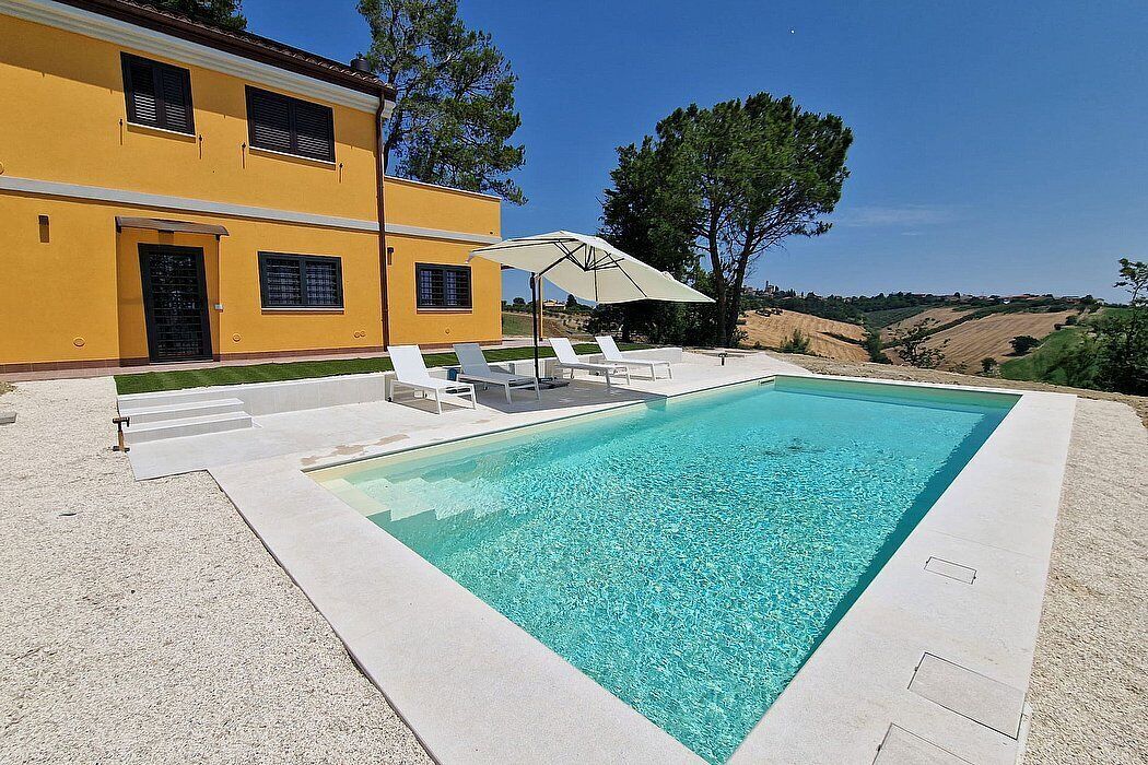 A rectangular swimming pool with clear blue water, surrounded by a white deck. There are white lounge chairs and a large umbrella providing shade. The pool is adjacent to a yellow building with multiple windows and a door.