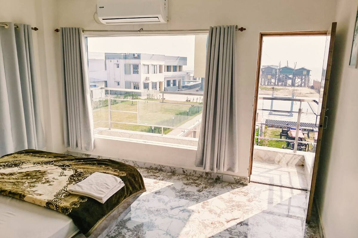 A hotel room with a bed, an air conditioner, a large window with curtains, and a view of buildings and a body of water.