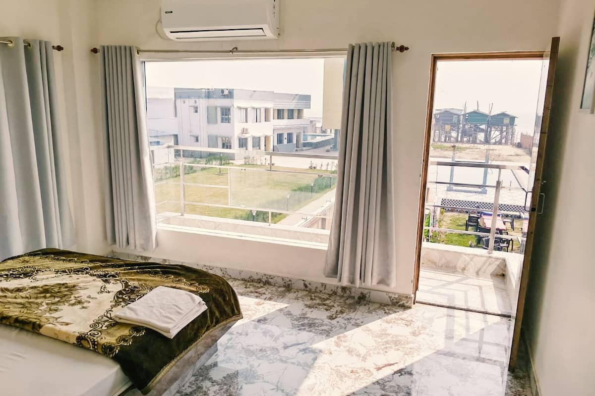 A hotel room with a bed, an air conditioner, a large window with curtains, and a view of buildings and a body of water.