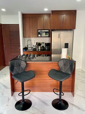 Una cucina moderna con mobili in legno, un frigorifero in acciaio inossidabile, un forno a microonde e una macchina da caffè. Sono presenti due sgabelli neri e un piano di lavoro in marmo.