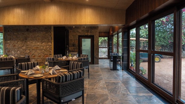 A restaurant with wicker furniture, stone walls, and large windows overlooking a garden. - The Farm Stay by Aamaghati (Sawai Madhopur)