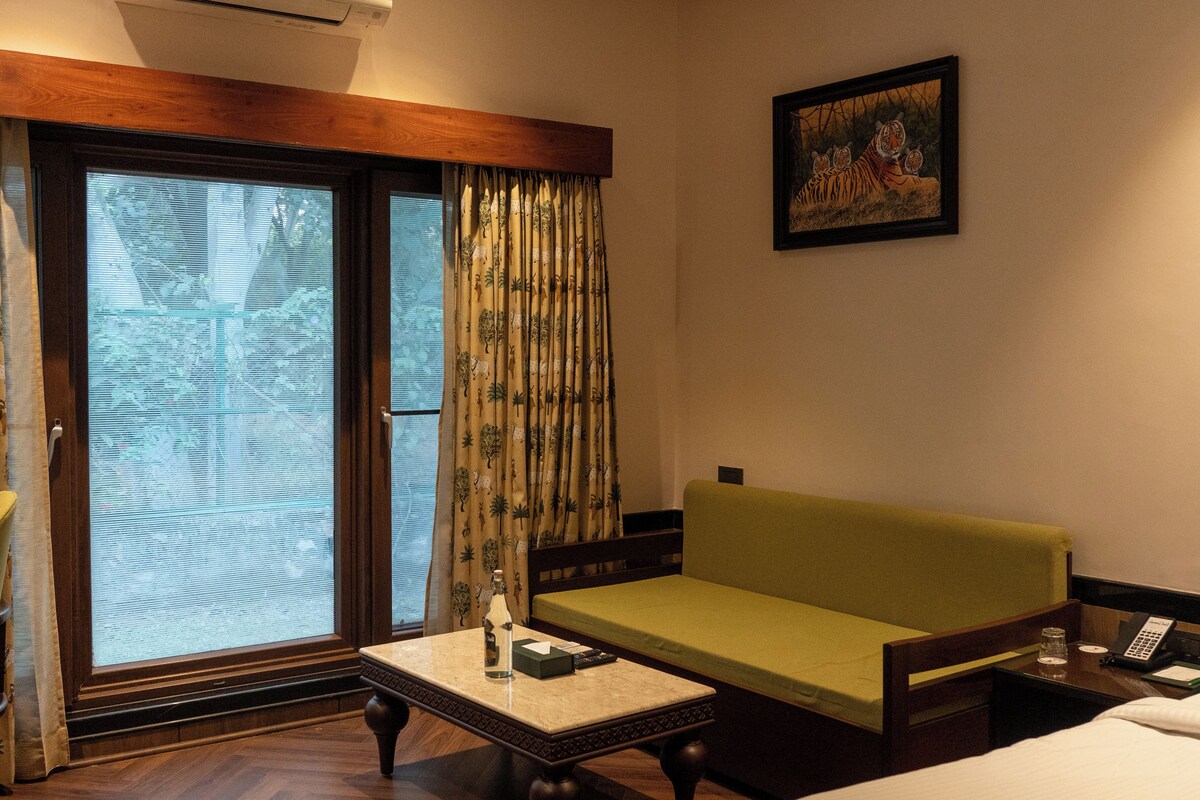 A hotel room with a wooden floor, a green sofa, a small table with a bottle and a glass, a window with a view of trees, and a painting on the wall.
