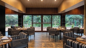 A restaurant with wicker furniture, large windows, and a view of greenery. - The Farm Stay by Aamaghati (Sawai Madhopur)