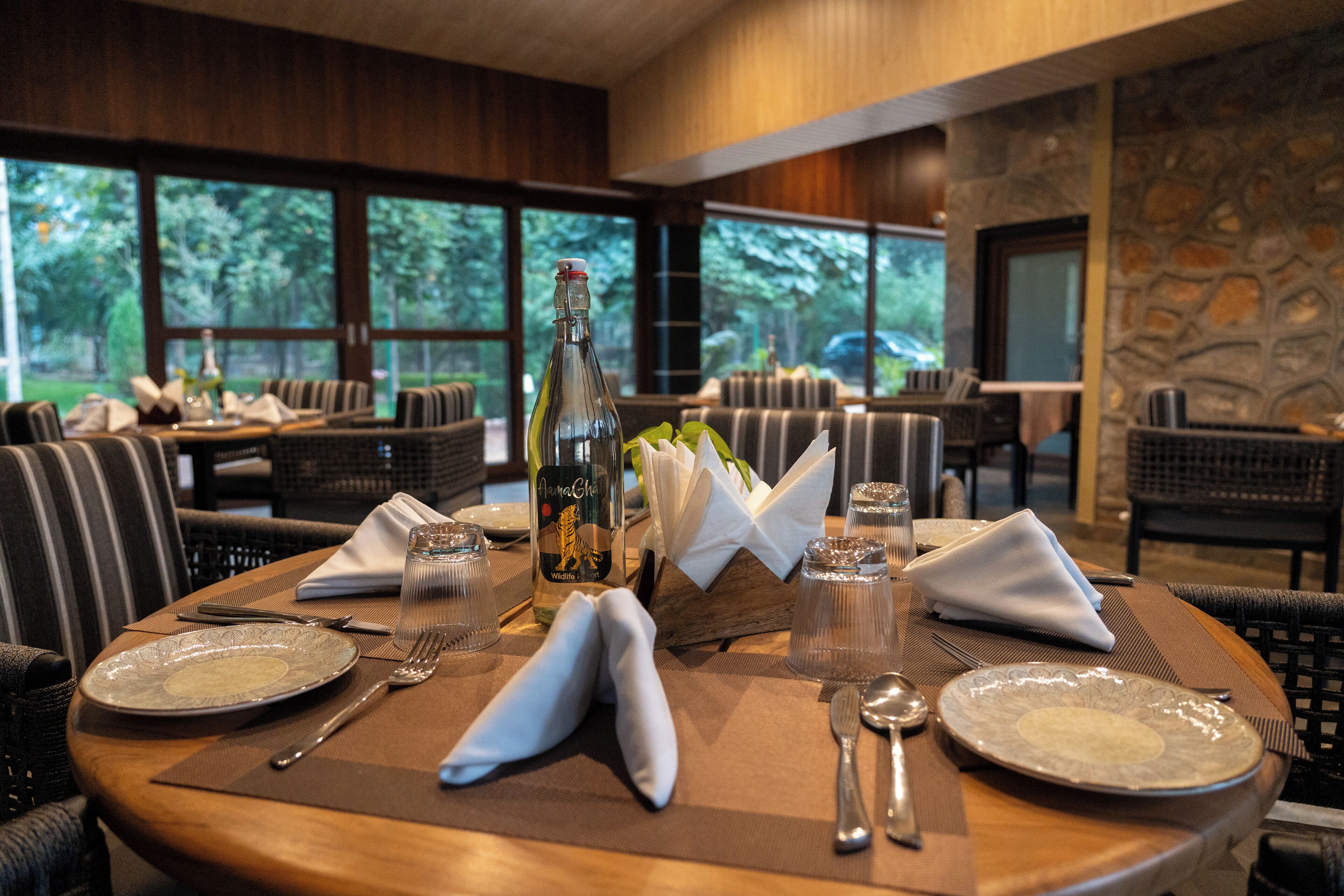 A dining table set with plates, cutlery, a bottle, and glasses, with a view of outdoor seating and greenery through large windows.