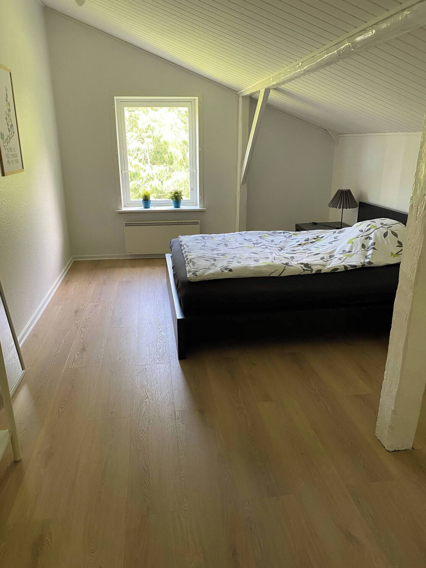 Une chambre à coucher avec un lit, une fenêtre ornée de plantes, une table de chevet et une lampe.