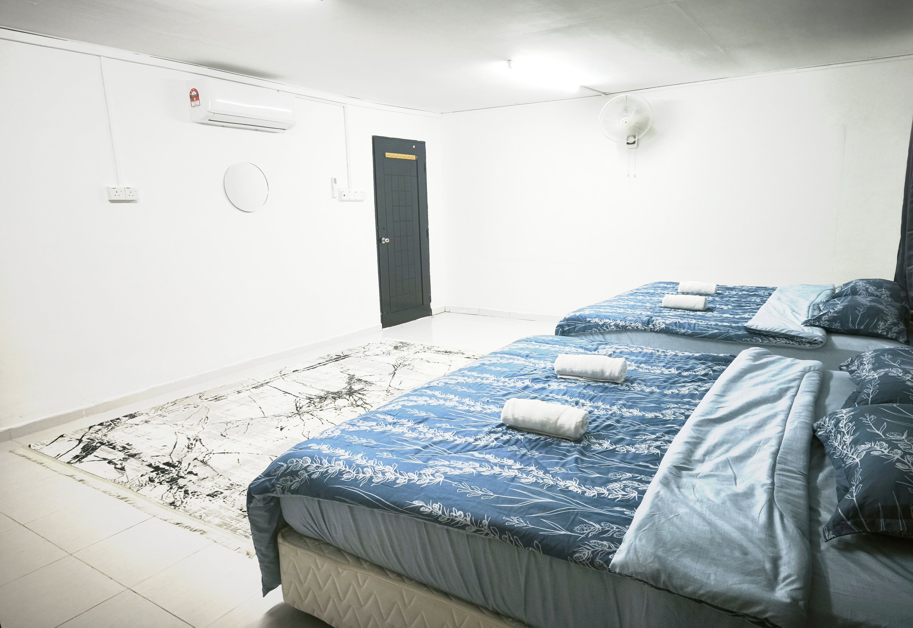 A hotel room with a bed, a bedside table, a wall-mounted air conditioner, a door, and a light switch.