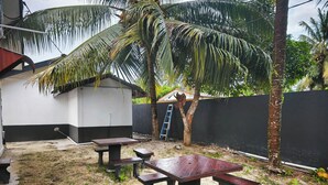 Un espace extérieur aménagé avec des tables de pique-nique et des bancs, entouré d’un petit muret et de palmiers.