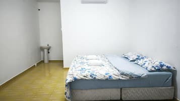 Une petite chambre avec un lit simple, disposant d’un aménagement simple : un lit, une petite table de chevet, un climatiseur fixé au mur et des toilettes.
