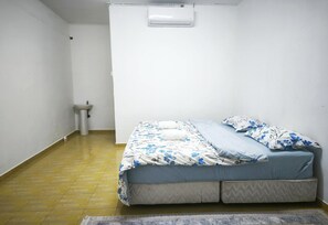 Egy kis, egyágyas szoba egyszerű berendezéssel, amely tartalmaz egy ágyat, egy kis éjjelit, egy falra szerelt légkondicionálót és egy mosdót.
