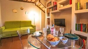 A living room with a green sofa, a glass coffee table with a bowl of pastries, wine glasses, and a flat-screen TV.