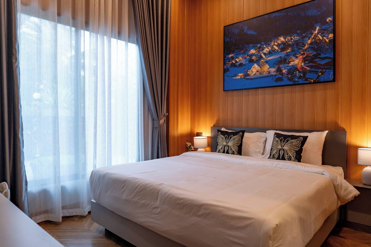 Habitación de hotel con una cama grande, una mesita de noche, una lámpara y un cuadro de un pueblo cubierto de nieve en la pared.