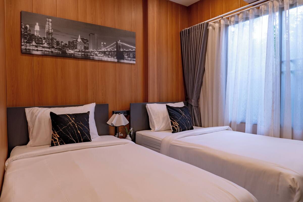 Habitación de hotel con dos camas, un cuadro de paisaje urbano, una lámpara y una ventana con cortinas.