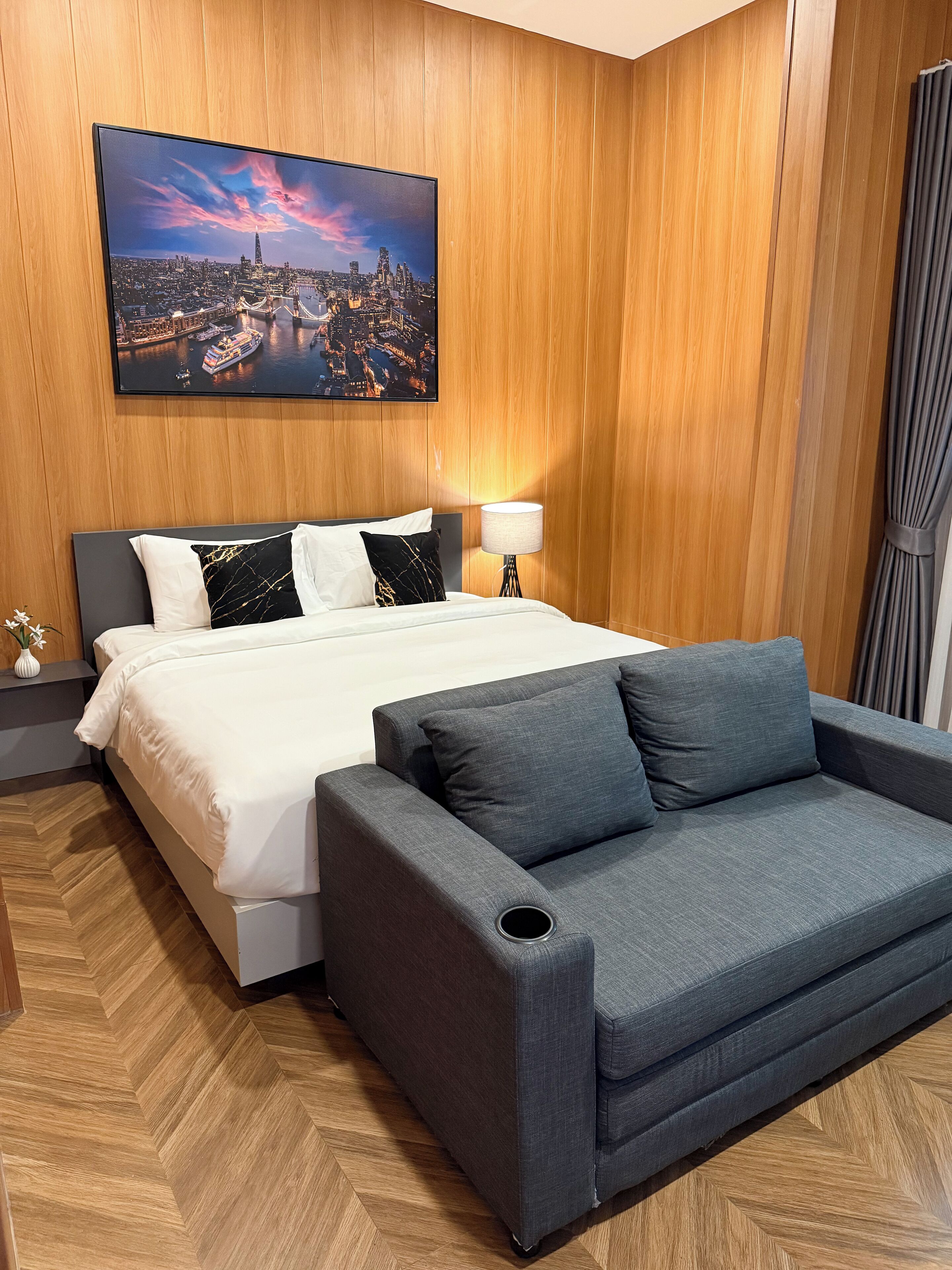 A hotel room with a bed, a gray sofa, a nightstand, a lamp, and a cityscape painting on the wall.