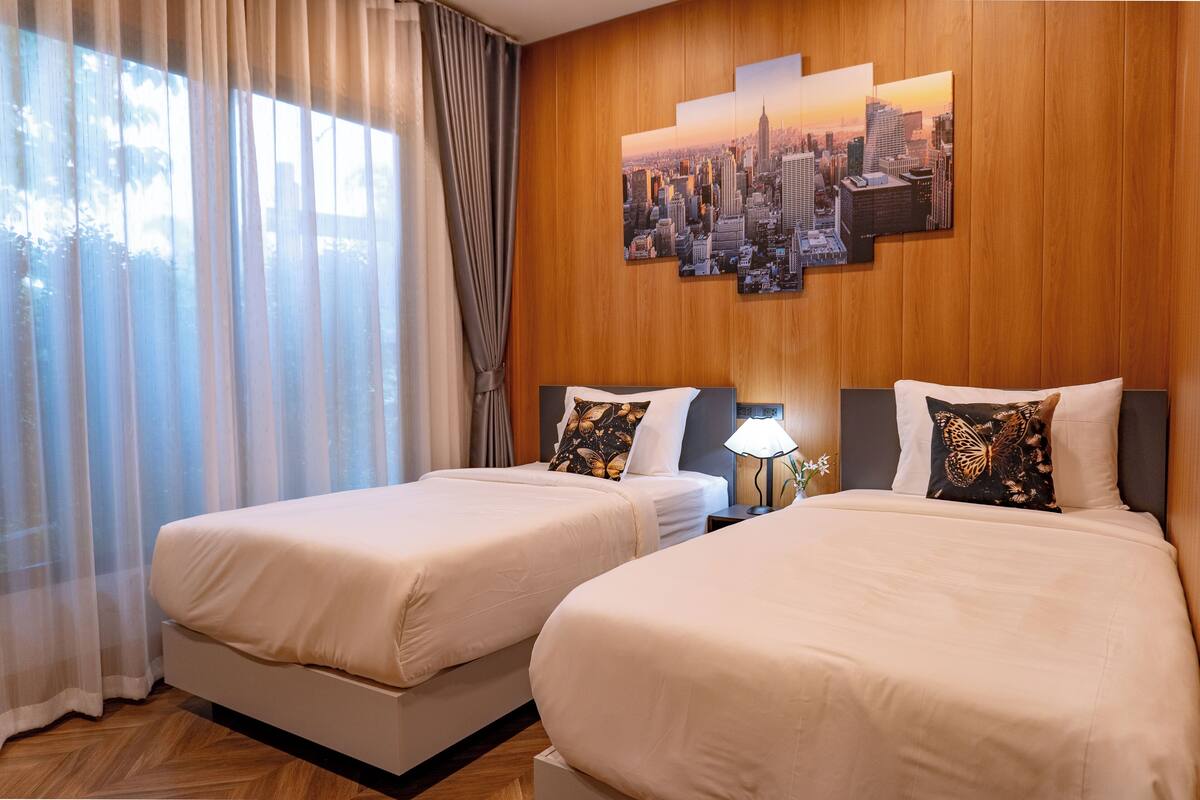 Habitación de hotel con dos camas, un cuadro de arte urbano en la pared, una mesita de noche con lámpara y cortinas translúcidas.