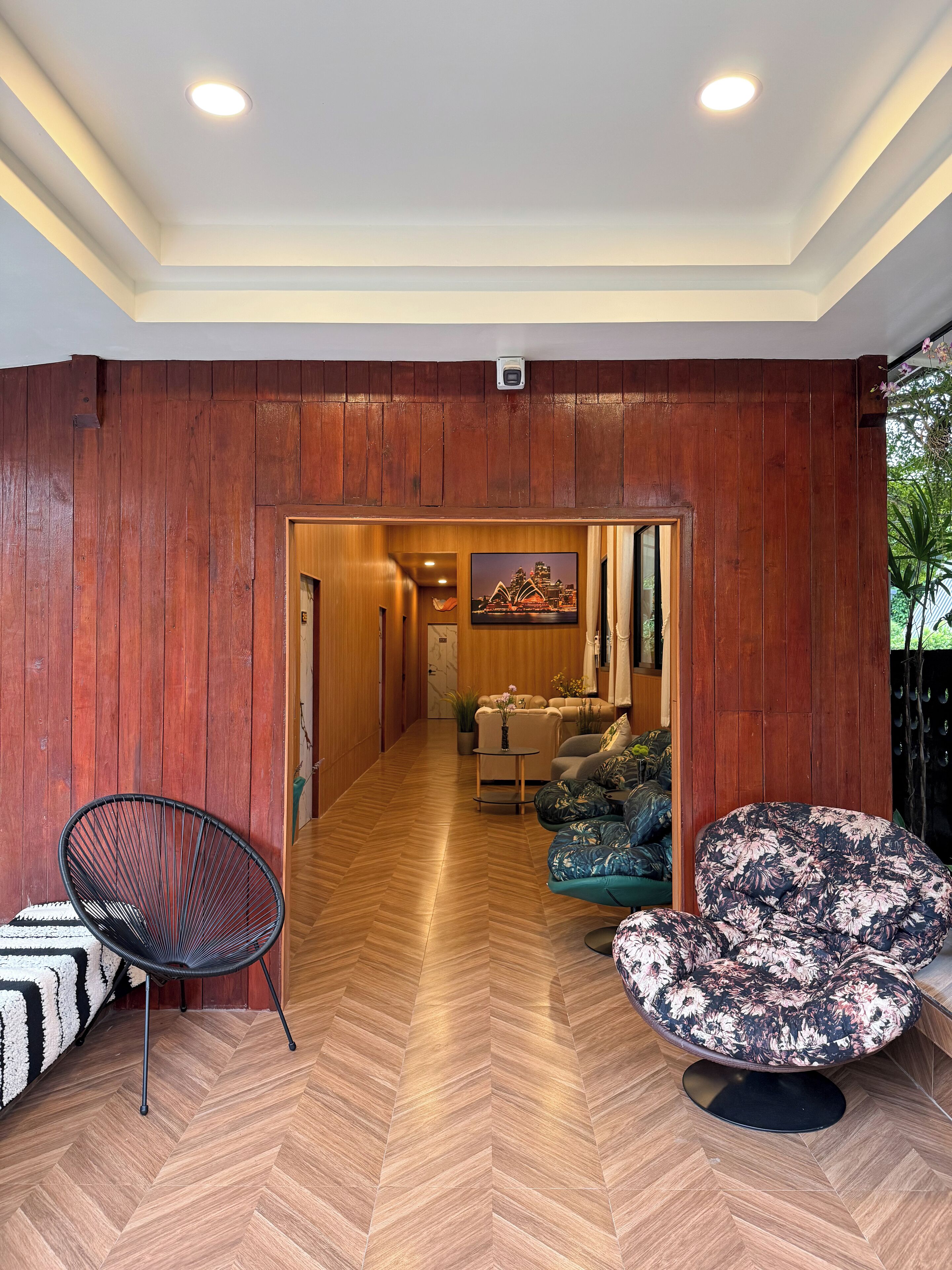 A hallway with wooden paneling, a black wire chair, a floral sofa, and a view into a living area with a sofa and a painting of a building.