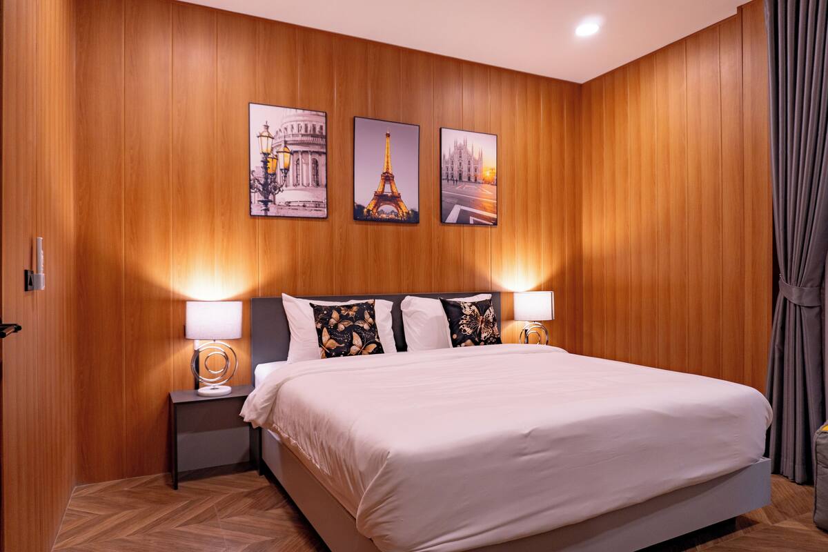 Habitación de hotel con pared de paneles de madera, una cama grande con ropa blanca, dos lámparas de noche y tres cuadros con imágenes de monumentos de París.