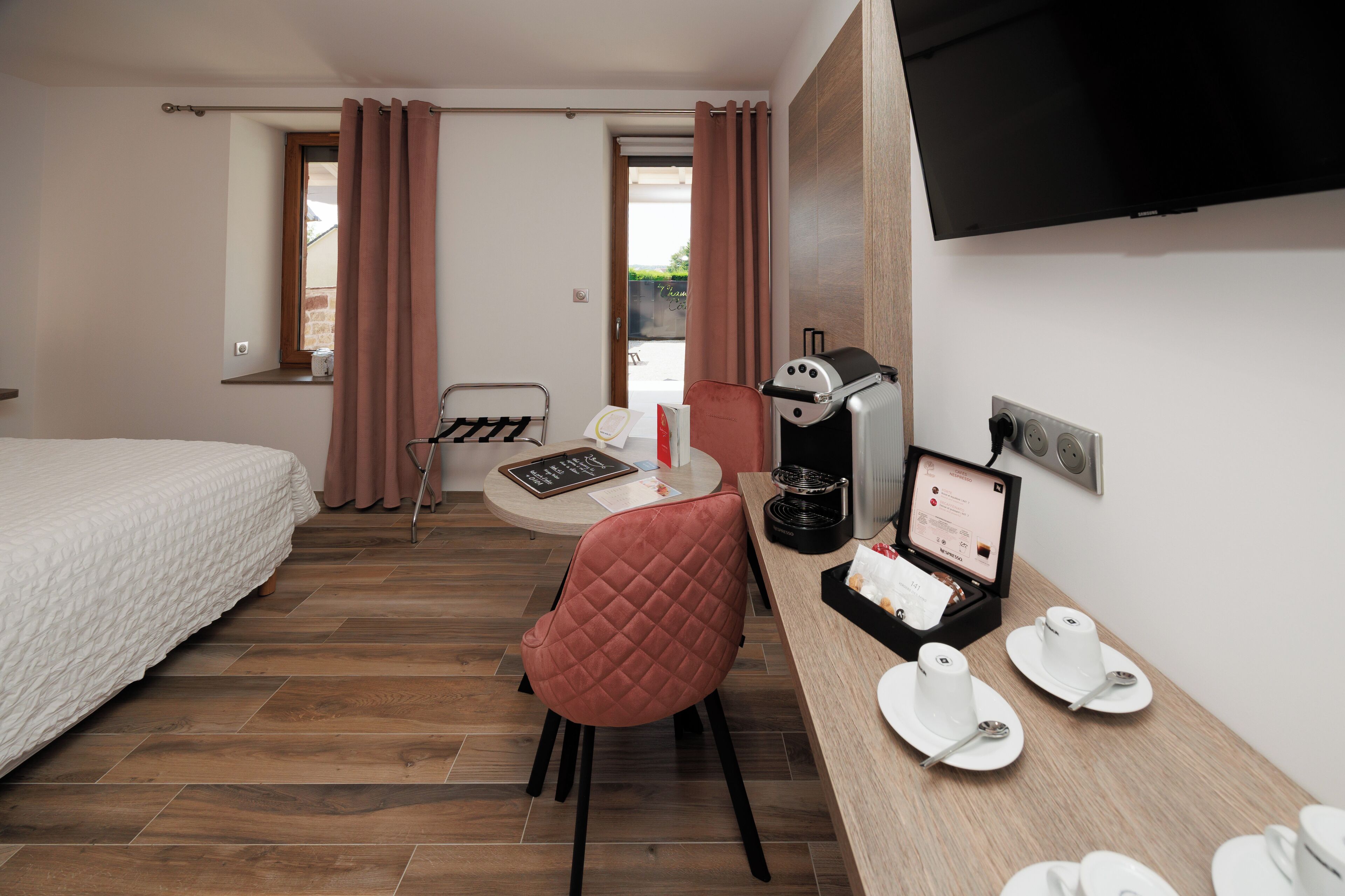 A hotel room with a bed, a desk with a computer, a chair, a coffee machine, and a TV.