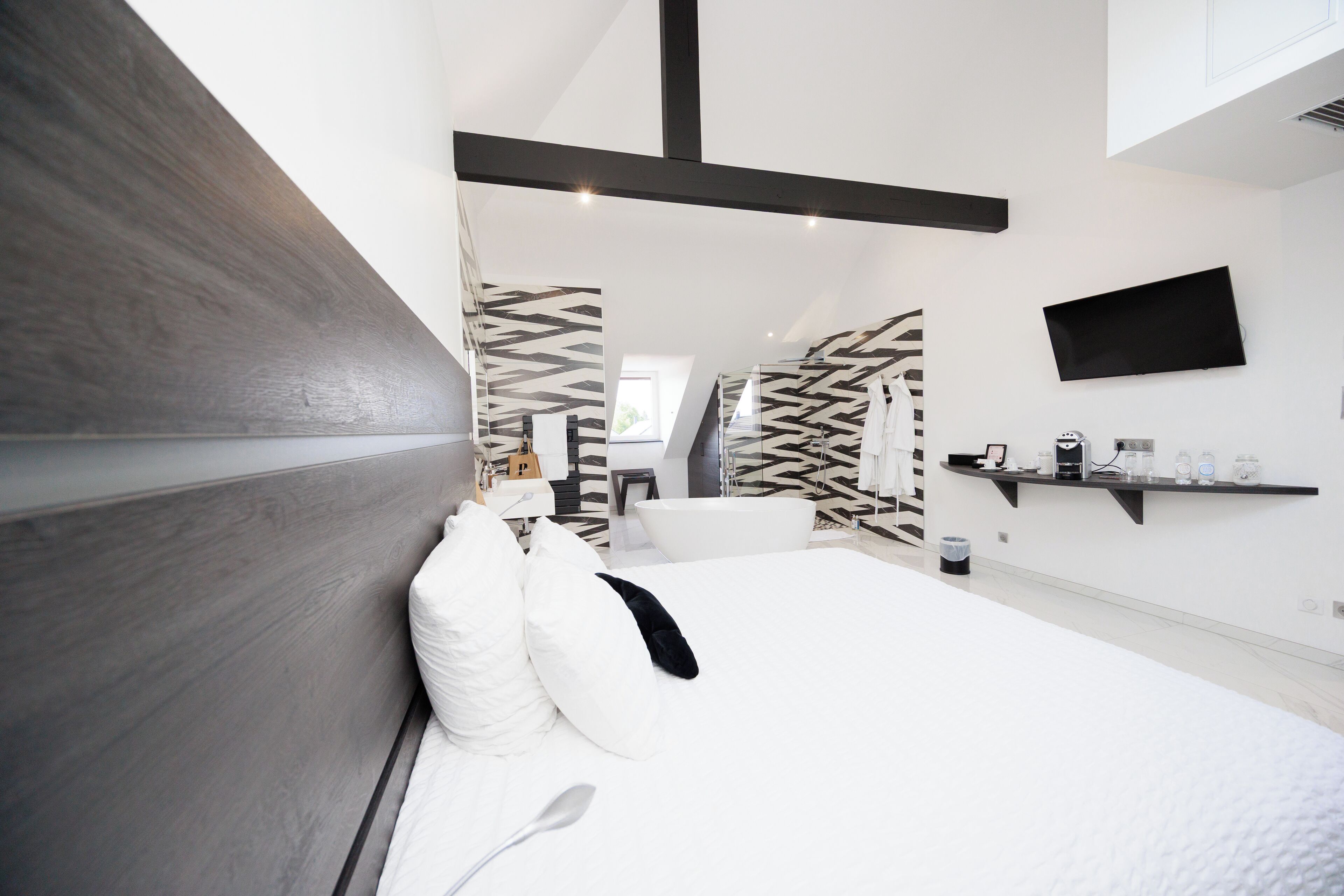 Un dormitorio moderno con una cama grande, un banco empotrado, un televisor montado en la pared y un baño con un revestimiento de pared a cuadrícula blanco y negro.