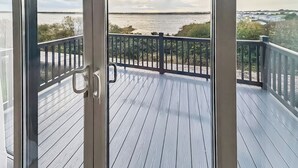 Une terrasse en bois dotée d’une rambarde, offrant une vue sur une étendue d’eau et sur des bâtiments situés au loin.