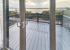 A wooden deck with a railing, overlooking a body of water and distant buildings. - Superior 2 Bed Lodge (14ft wide) (Haverigg, Millom)