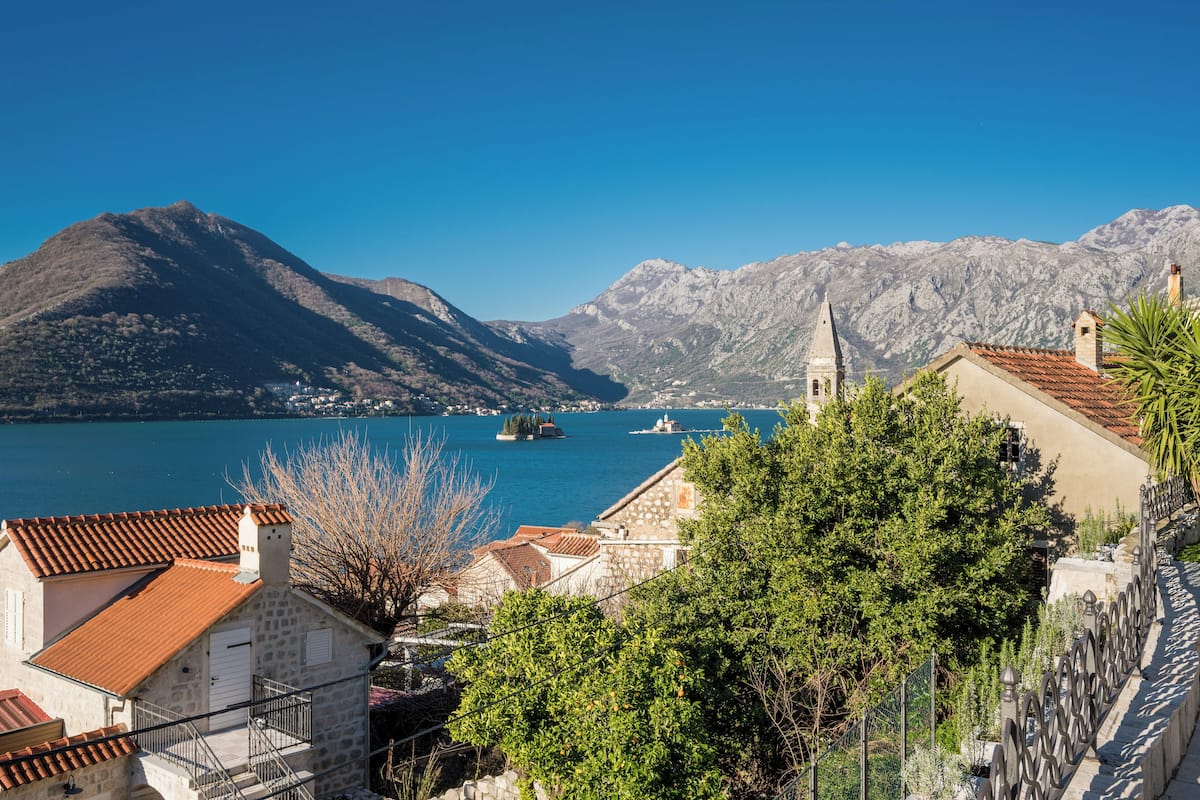 A coastal town with stone buildings, a church, and a clear view of the sea and mountains.