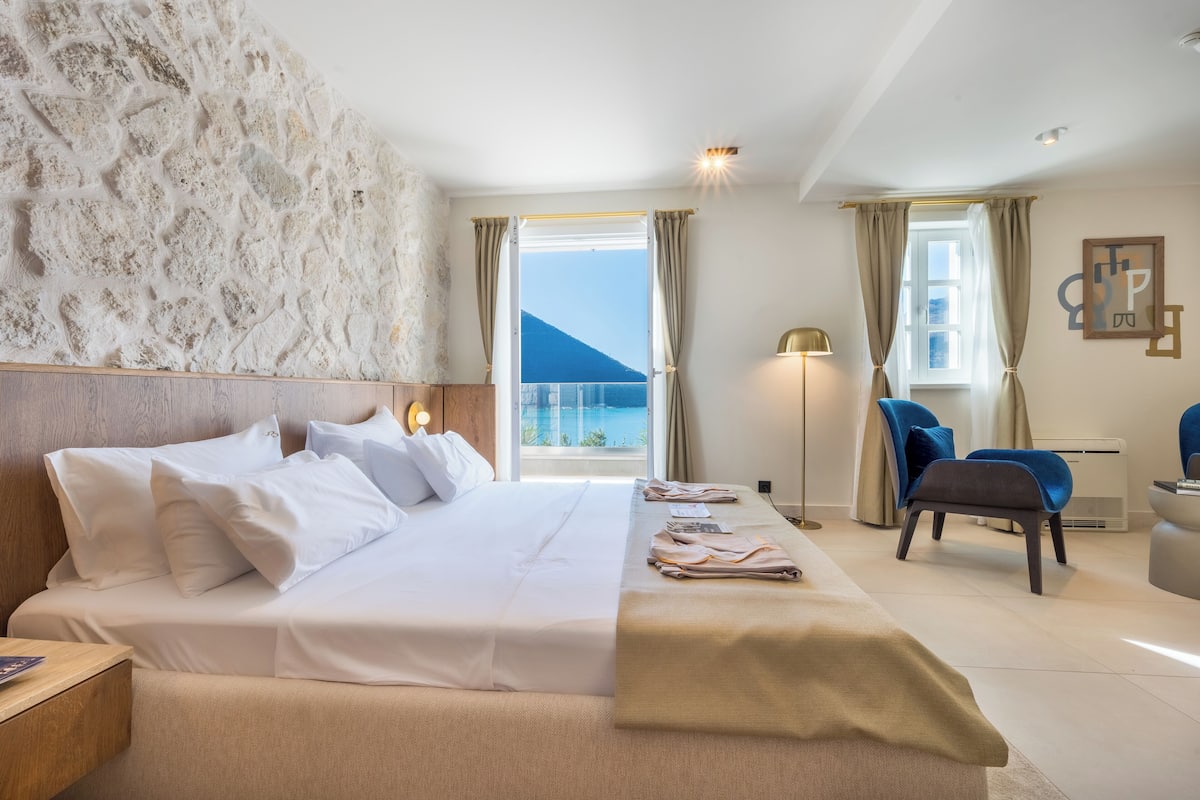 A hotel room with a large bed, a desk, a chair, a lamp, and a view of the sea.