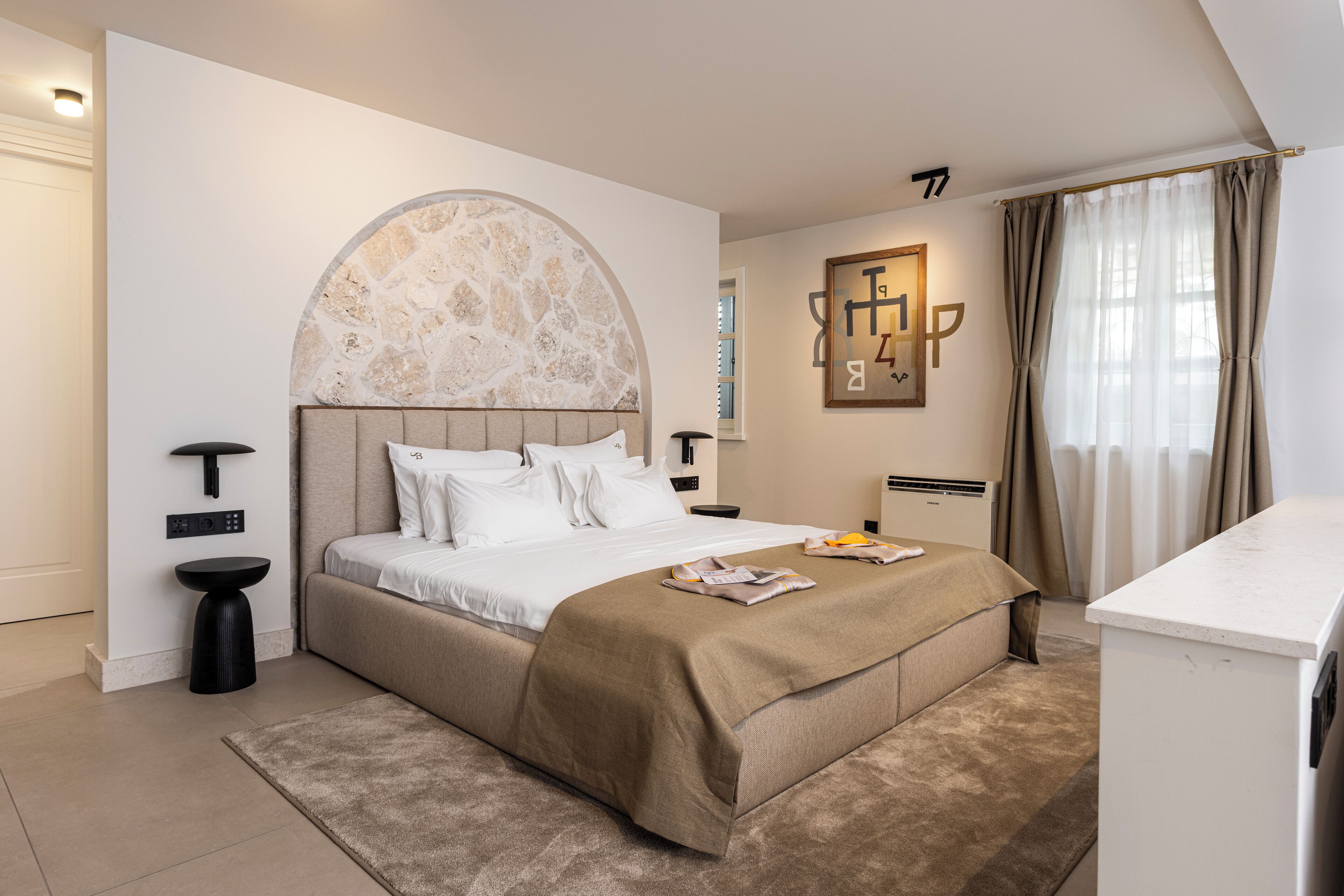 A modern bedroom with a large bed, a round stone accent wall, a bedside table, a small stool, and a built-in desk.