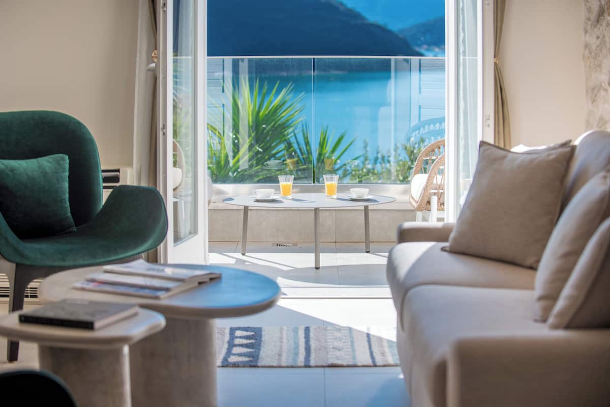 A modern living room with a green chair, a beige sofa, and a small round table with books. A balcony with a glass door leads to a scenic view of a body of water and mountains.
