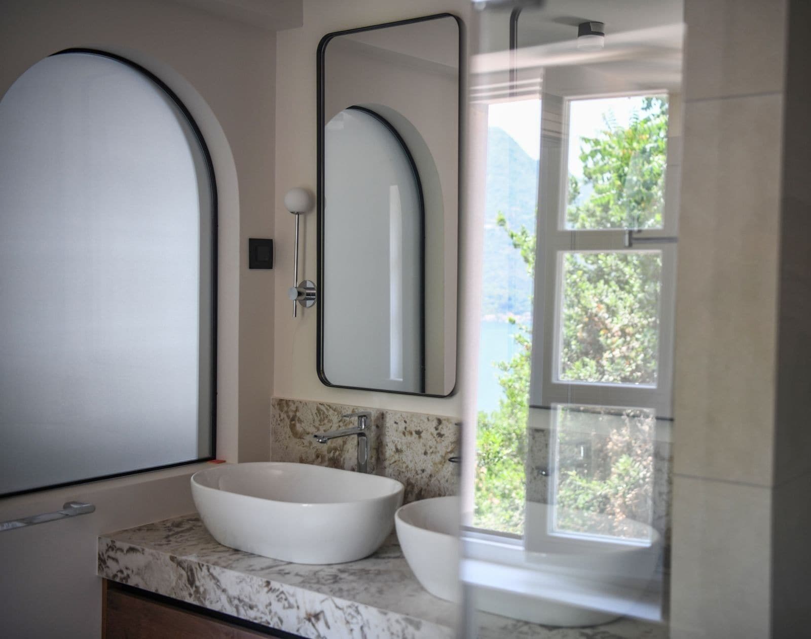 A bathroom with a large oval sink, a wall-mounted mirror, and a window with a view of greenery.
