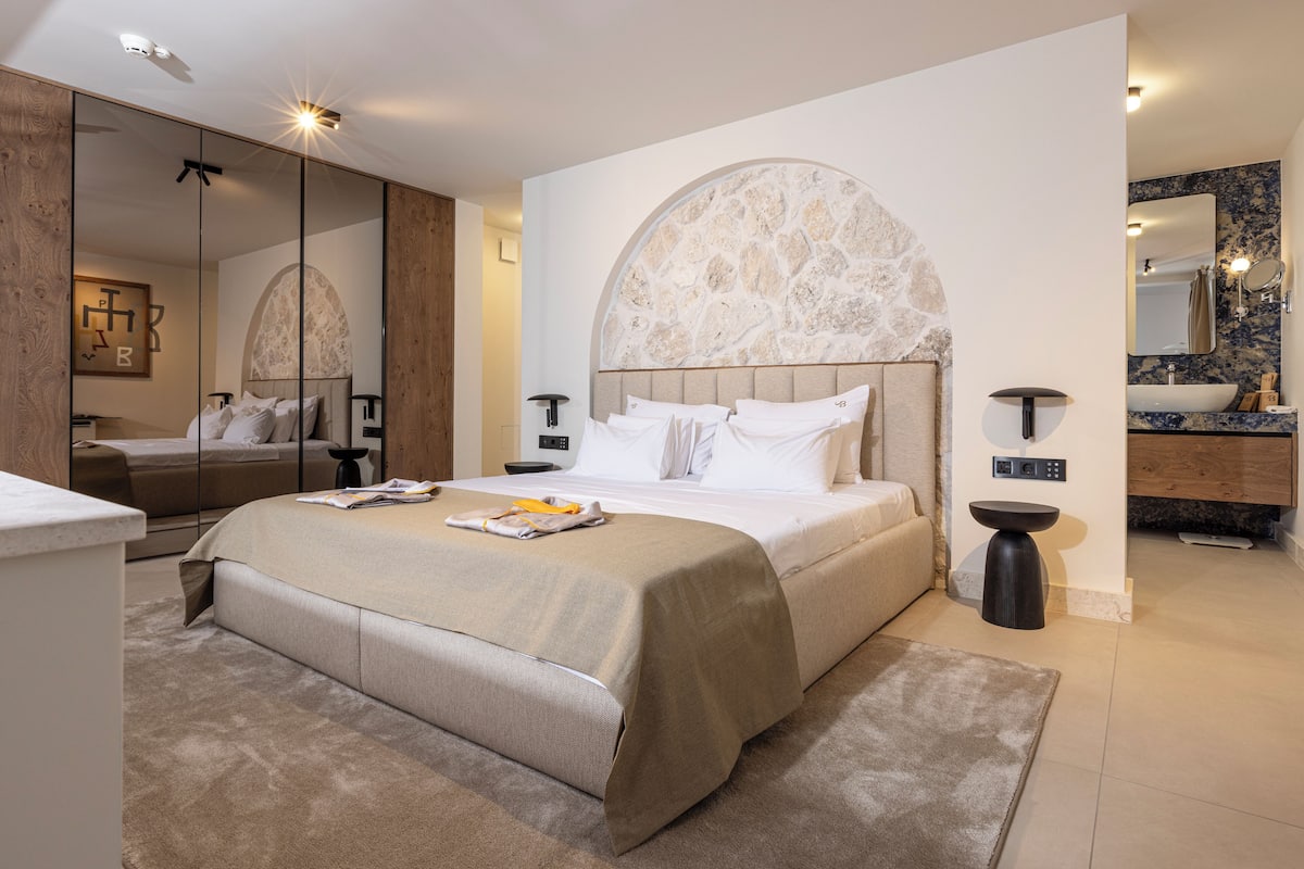A modern hotel room with a large bed, a vanity with a mirror, and a stone accent wall.