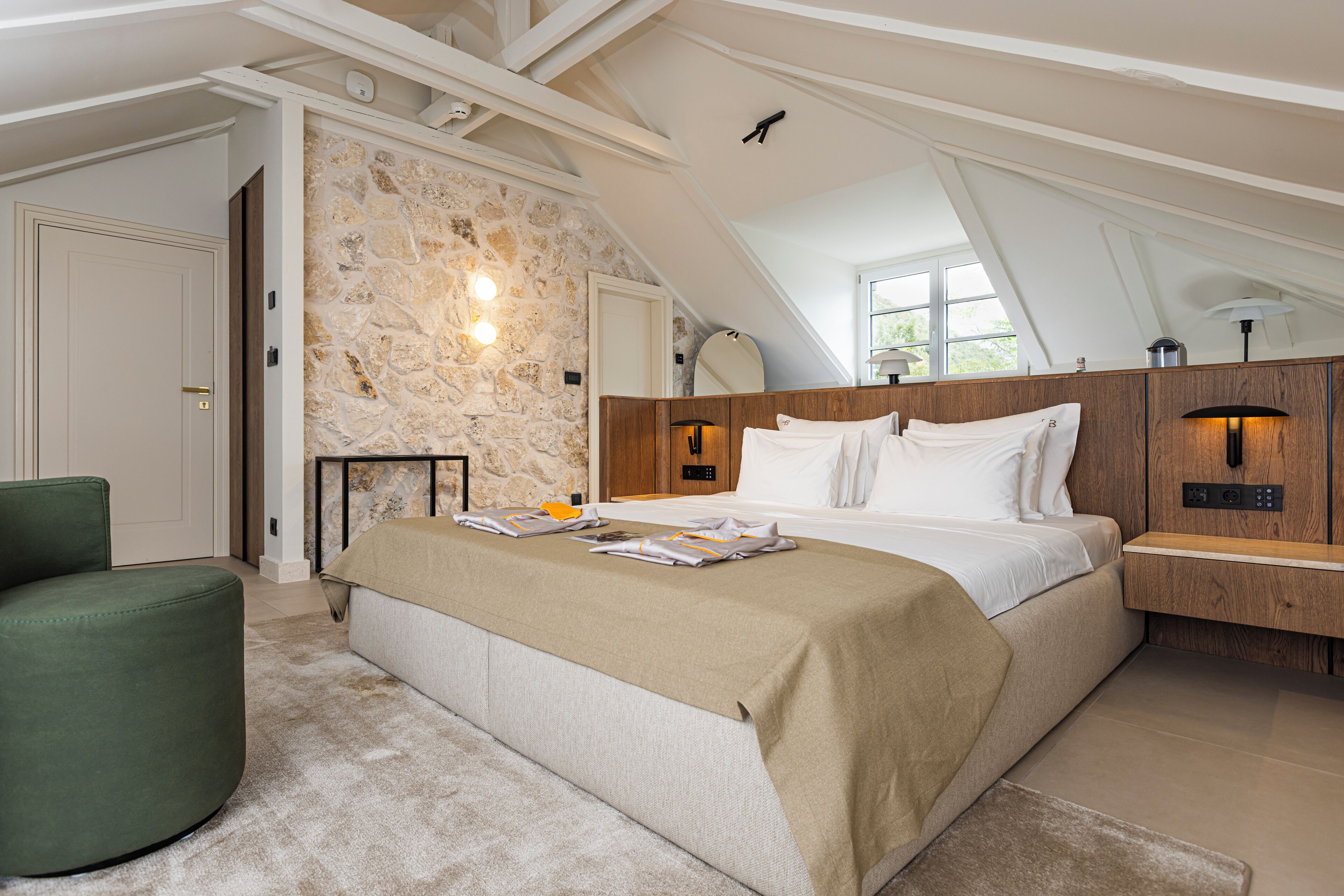 Un dormitorio con pared de piedra, cama con ropa blanca, sillón verde y mesita de noche de madera con una lámpara.