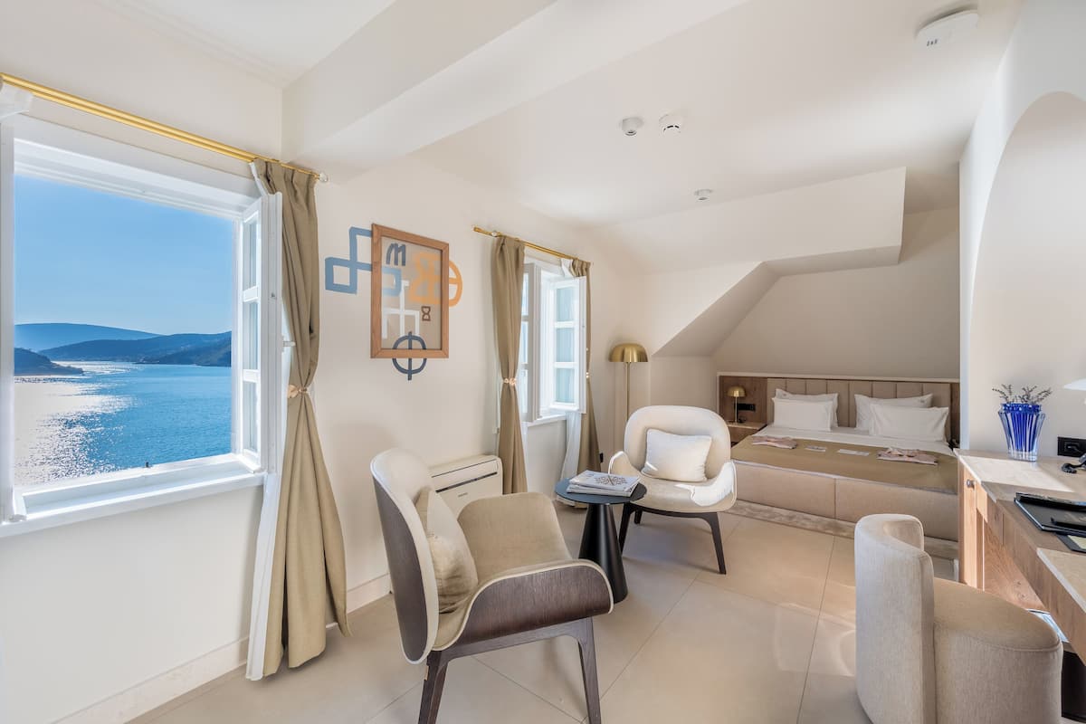 A modern hotel room with a large window offering a sea view, a bed with white linens, a desk with a lamp, and a chair.