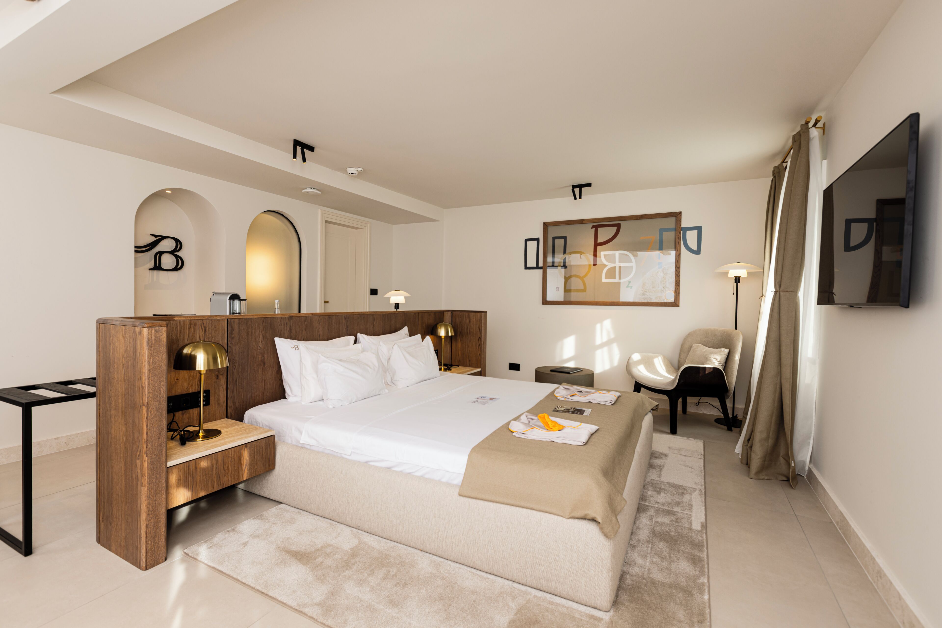 Una habitación de hotel moderna con una cama grande, cabecera de madera, mesitas de noche con lámparas, un televisor de pantalla plana, una silla y un espejo en la pared.