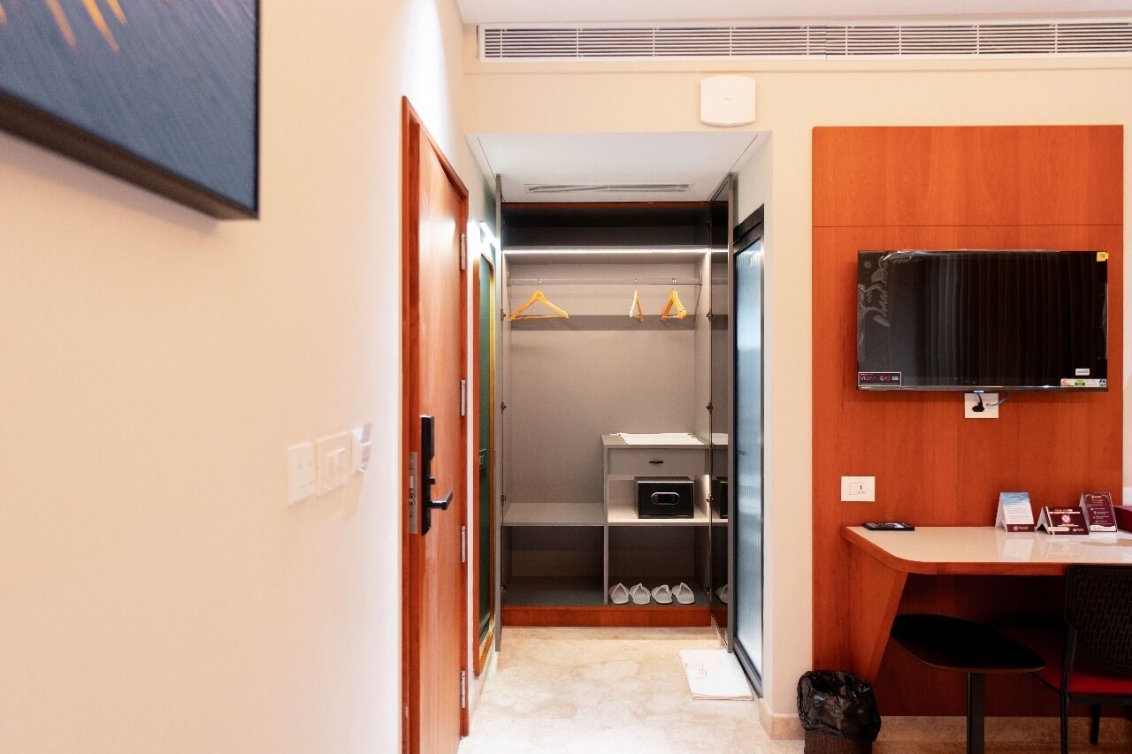 Habitación de hotel con escritorio de madera, televisor montado en la pared, armario con ropa colgada y zapatero.