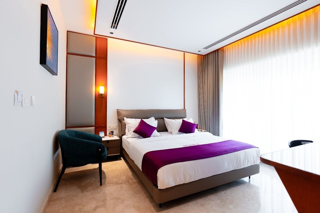 Une chambre d’hôtel moderne, dotée d’un grand lit, d’une table de chevet, d’une chaise et offrant une vue sur la ville grâce à de grandes fenêtres.