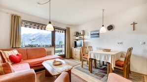 Una sala de estar acogedora con un sofá, una mesa de comedor y sillas. Se ve un televisor y una estantería al fondo.