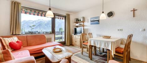 A cozy living room with a sofa, a dining table, and chairs. There is a TV and a bookshelf in the background.