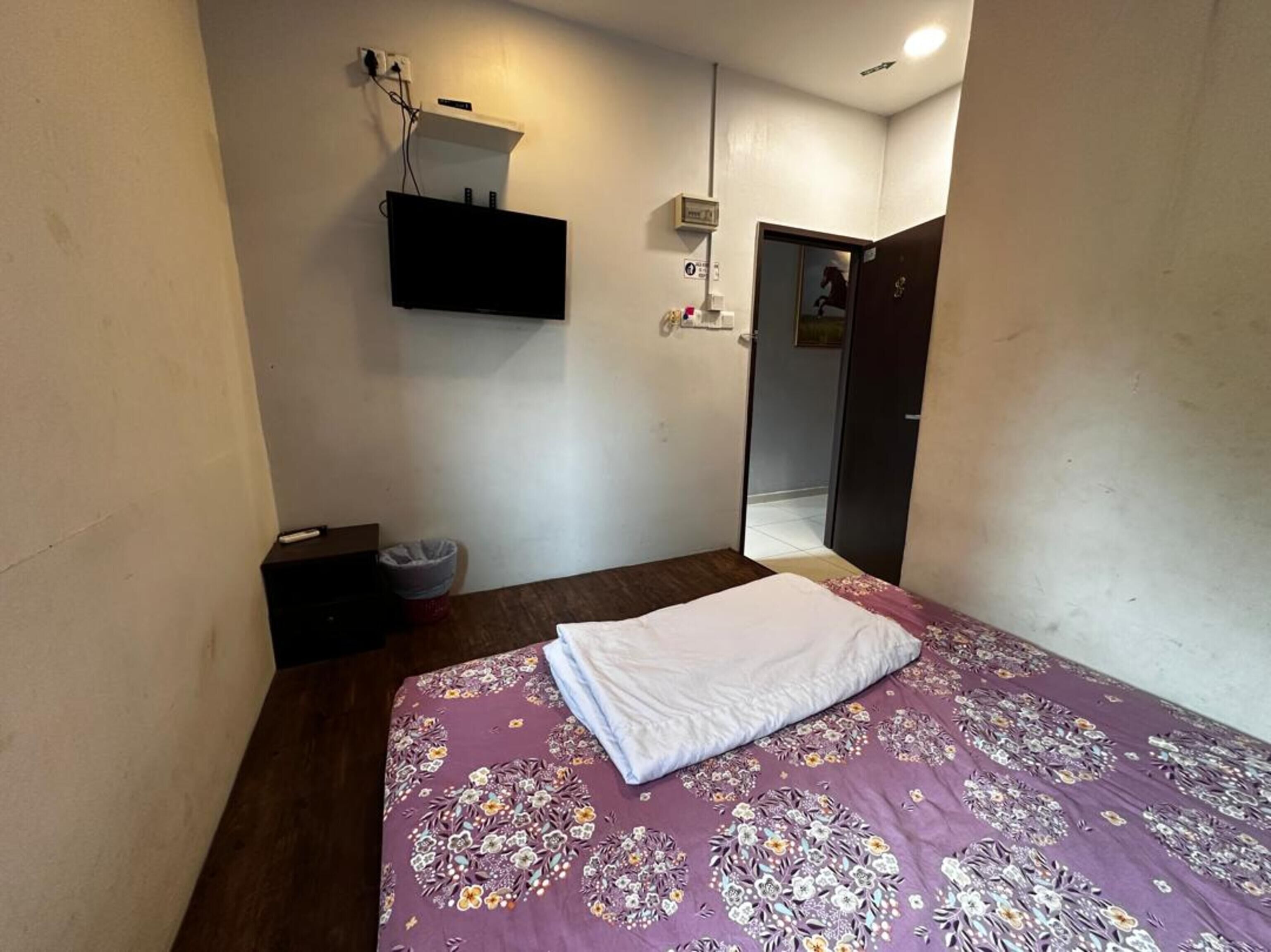 A bedroom with a bed, a small bedside table, a wall-mounted TV, and a door leading to another room.
