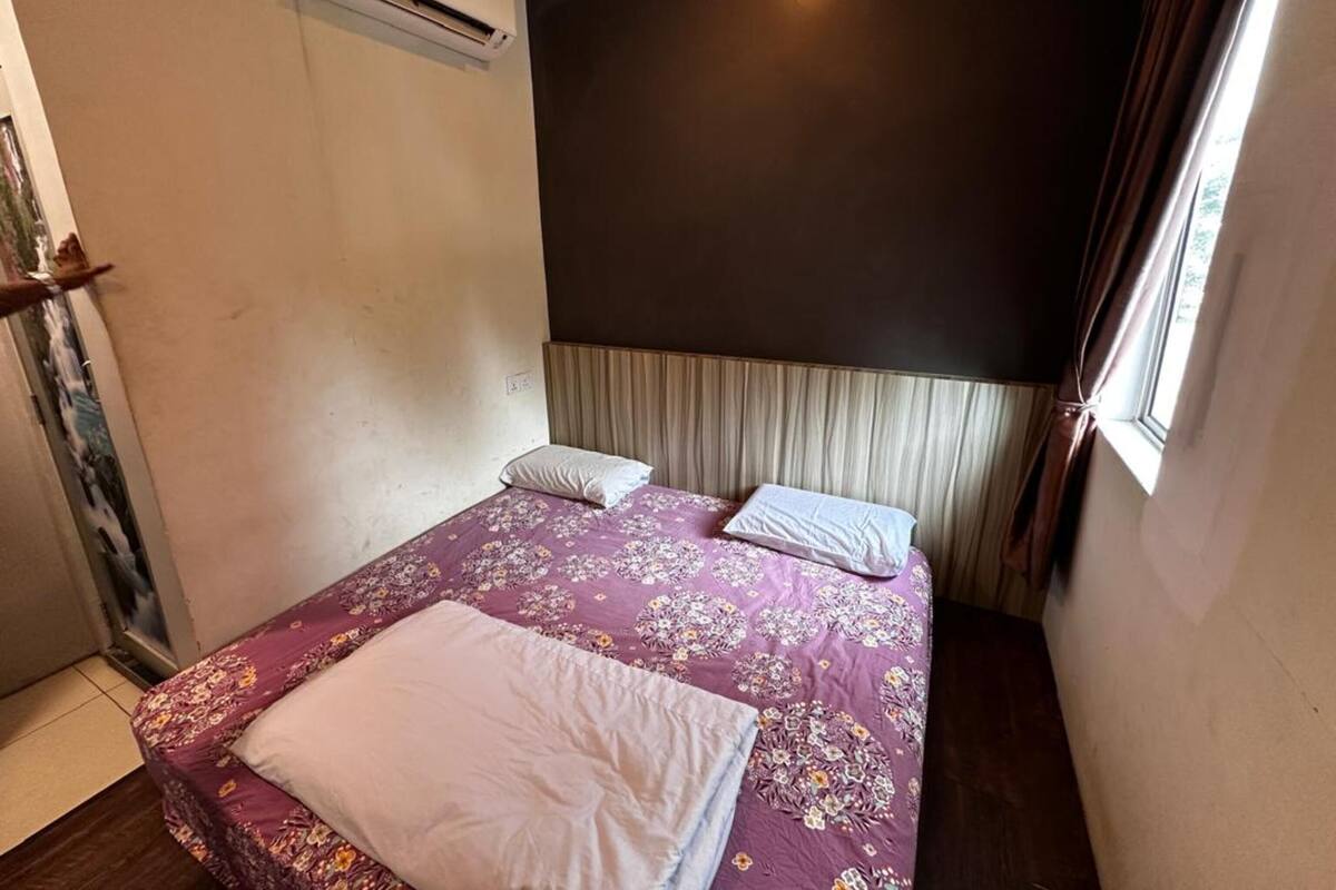 A small, single-bed room with a dark headboard, a window with curtains, and an air conditioning unit on the wall.
