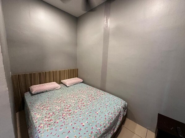 A small, single-bed room with a wooden headboard, a floral bedspread, and a plain grey wall.