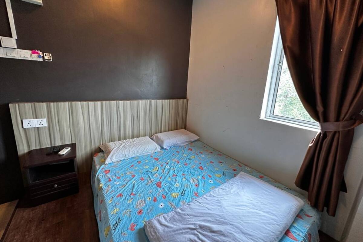 A single bed with a floral bedspread, a bedside table with a remote control, and a window with brown curtains.