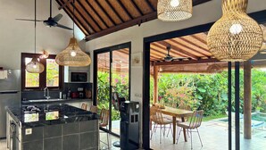 Une cuisine moderne dotée d’un îlot en marbre noir, d’un four à micro-ondes encastré et d’une machine à café. On y trouve également un coin repas avec une table et des chaises en bois, et une vue sur un jardin luxuriant grâce à de grandes portes vitrées.
