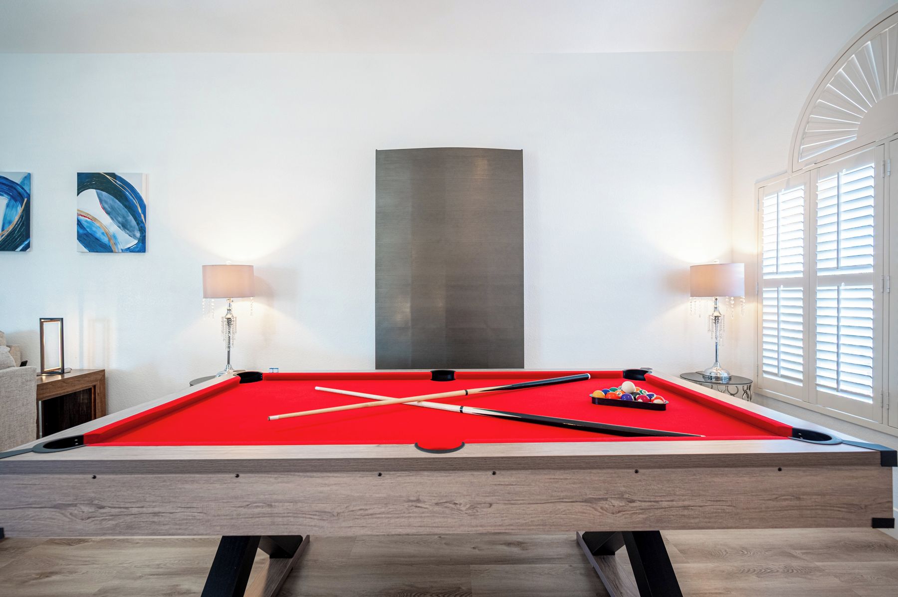A pool table with a red felt surface, surrounded by a wooden frame, in a room with white walls and a large abstract painting.