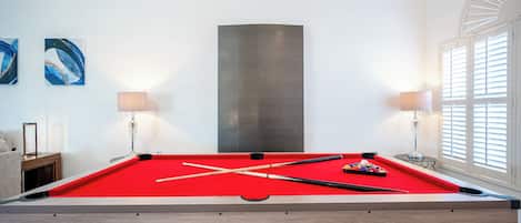 A pool table with a red felt surface, surrounded by a wooden frame, in a room with white walls and a large abstract painting.