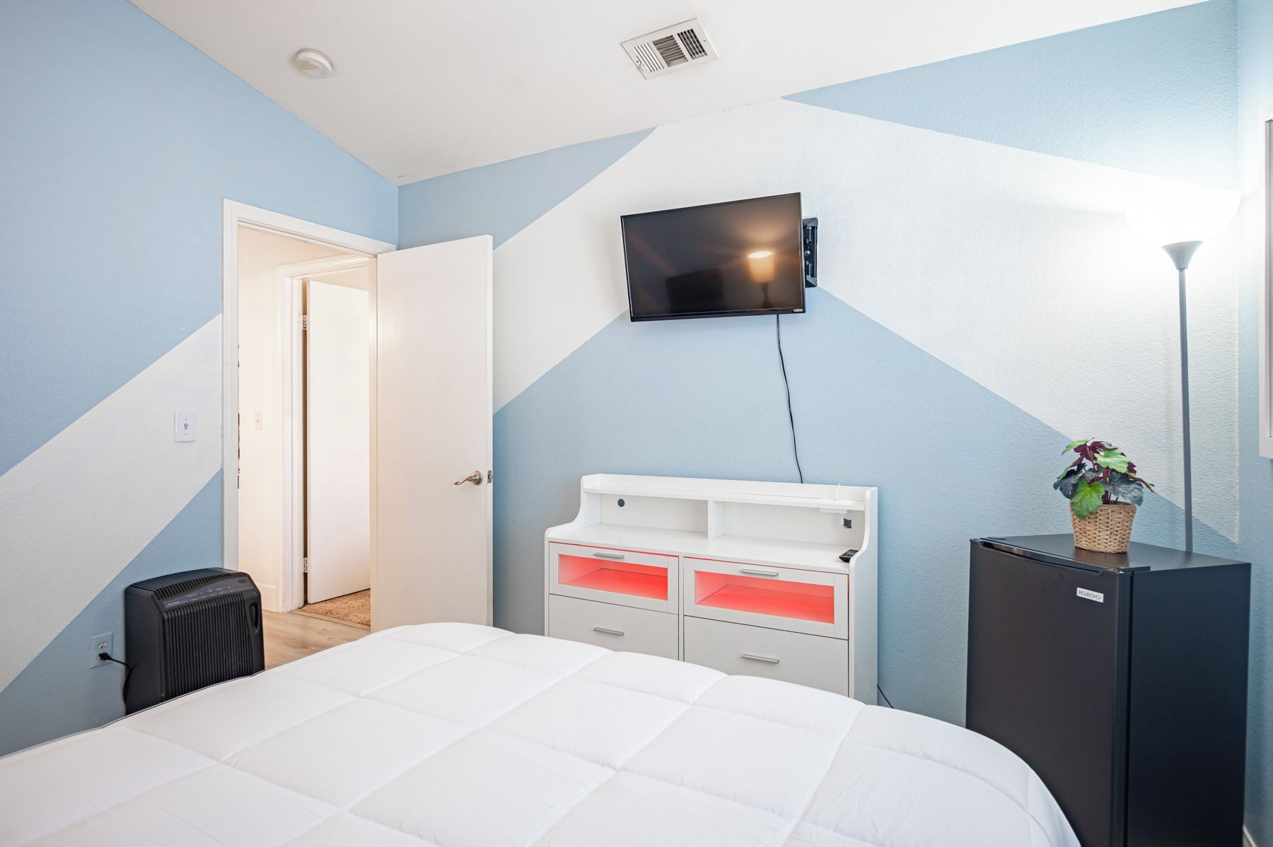 Een slaapkamer met een bed, een aan de muur bevestigde televisie, een dressoir met lades, een radiator en een deur naar een andere kamer.