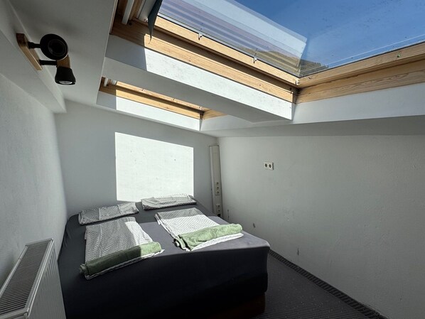 Quarto pequeno com cama, claraboia e radiador.