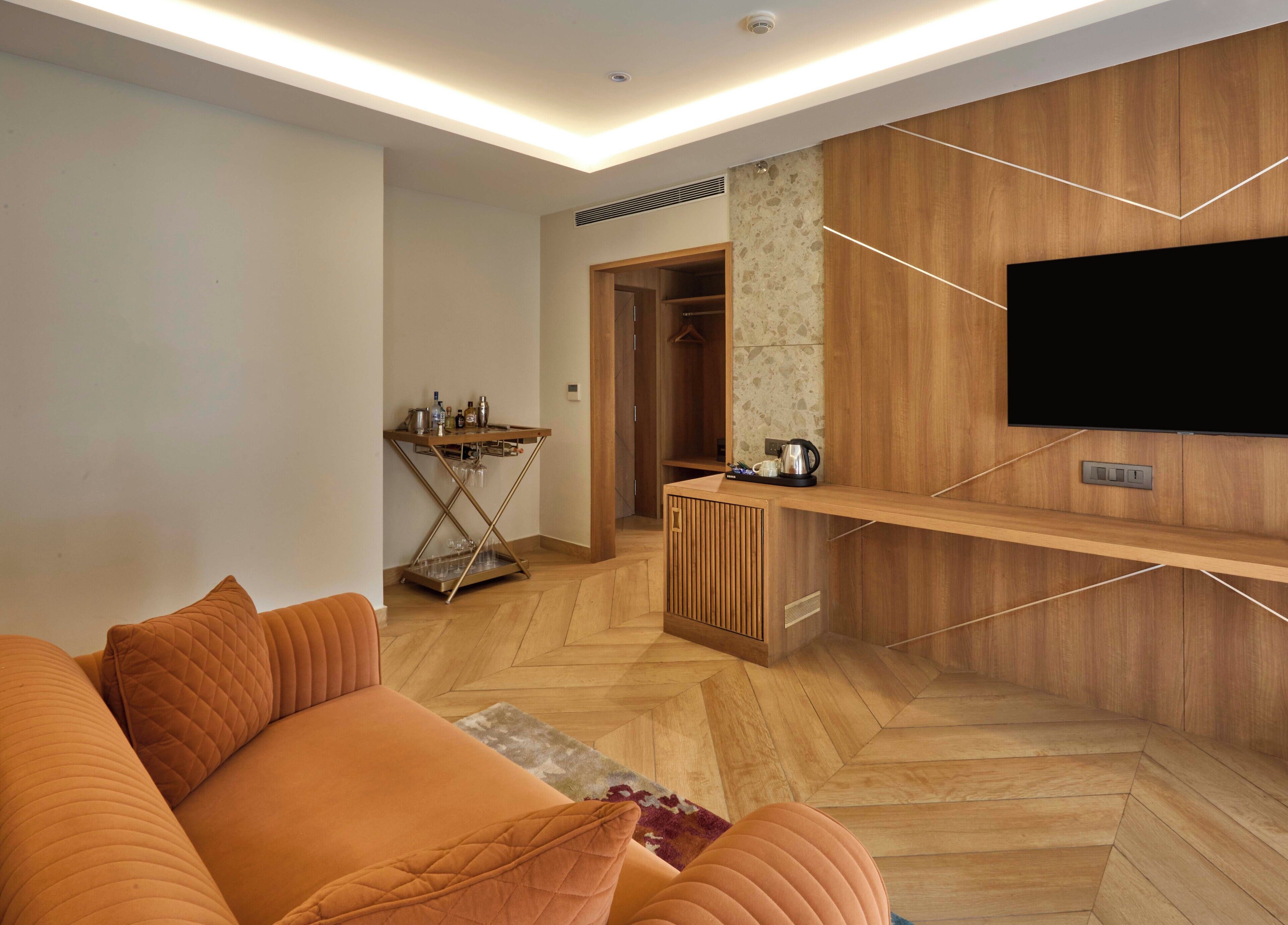 A modern living room with a wooden herringbone floor, a sofa with orange cushions, a coffee table, a TV mounted on a wooden wall, and a small kitchenette area with a coffee machine and wine glasses.
