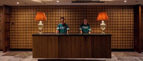 A reception desk with two staff members, wooden paneling on the wall, and two lamps.